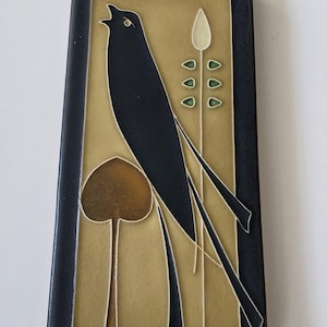 Puede incluir: Azulejo cerámico rectangular con un diseño de pájaro negro. El pájaro canta con el pico abierto, sobre un fondo dorado con elementos vegetales estilizados. El azulejo tiene un borde negro.