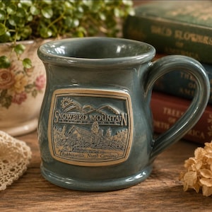 May include: A blue-grey ceramic mug featuring a raised rectangular emblem on the front. The emblem depicts a mountain range, a cabin, and the text "SNOWBIRD MOUNTAIN" above "Robbinsville, North Carolina". The mug has a wide rim and a curved handle.