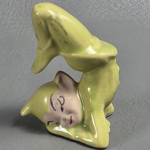 Pode incluir: Estatueta de cerâmica de um duende caprichoso em posição de parada de mãos. O duende é principalmente verde claro com um rosto rosa pálido, olhos pretos e boca vermelha. A estatueta tem um acabamento brilhante e é provavelmente um item decorativo.