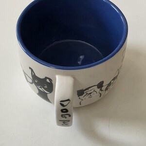 May include: White ceramic mug with a blue interior and a handle. The mug features black and white dog illustrations around the exterior. The handle has the words "Dog Mug" printed on it.