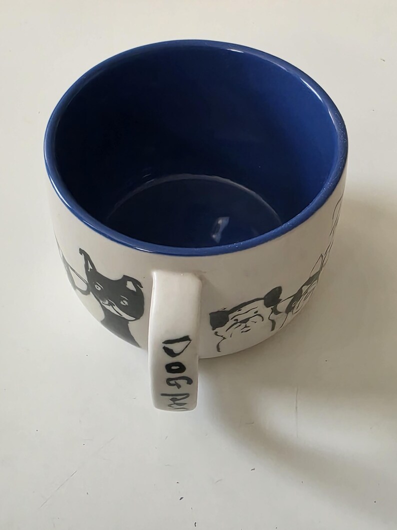 May include: White ceramic mug with a blue interior and a handle. The mug features black and white dog illustrations around the exterior. The handle has the words "Dog Mug" printed on it.