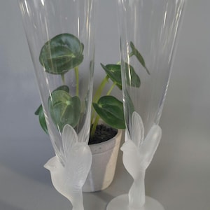May include: Two tall, clear glass champagne flutes featuring frosted glass stems shaped like birds with wings extended upward. The flutes have a smooth, flared rim and a circular base, showcasing a detailed, sculptural design on the stem.