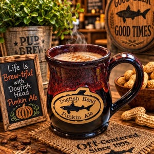 May include: A ceramic mug with a dark red and black glaze featuring a Dogfish Head Punkin Ale logo. The mug contains a steaming beverage and sits on a burlap coaster printed with the text Off-Centered Since 1995 and a shark silhouette.