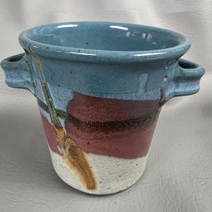 May include: Handmade ceramic vase with a blue rim and handles. The vase features bands of red, black, and white, with a unique abstract design. This decorative piece is perfect for holding flowers or as a standalone art object.