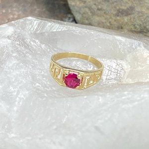May include: A gold ring with a red gemstone set in the center. The ring has a decorative design around the band.
