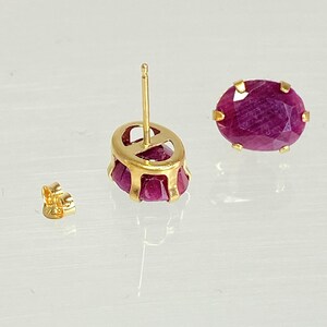 May include: A pair of gold earrings with oval-shaped ruby gemstones. The earrings have a post and butterfly back closure.