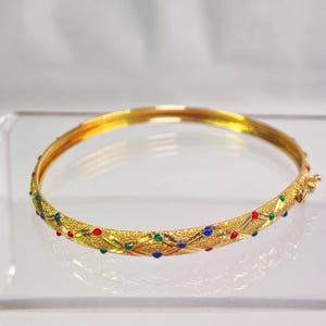 May include: A gold-tone hinged bangle bracelet featuring a textured surface with a cross-hatched pattern. The bracelet is decorated with small, inlaid round stones in vibrant red, green, and blue colors, and includes a secure metal clasp closure.