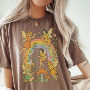 May include: Brown t-shirt featuring a whimsical design of fairies, a rainbow, and mushrooms. The fairies are depicted in various poses with colorful wings and dresses. The design is set against a backdrop of flowers and foliage.