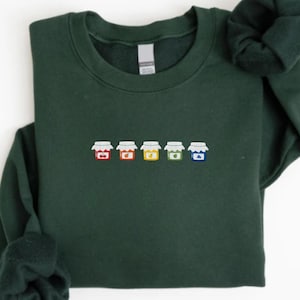 Op de afbeelding: Een donkergroene sweatshirt met een geborduurd ontwerp van vijf kleurrijke jampotten. De potten zijn rood, geel, oranje, groen en blauw, elk met een wit deksel. De sweatshirt heeft een ronde hals en lange mouwen.