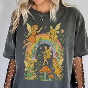 May include: Dark gray t-shirt featuring a whimsical illustration of fairies around a rainbow, mushrooms, and flowers. The fairies are depicted in yellow and orange dresses with butterfly wings. The shirt has a vintage, washed look.