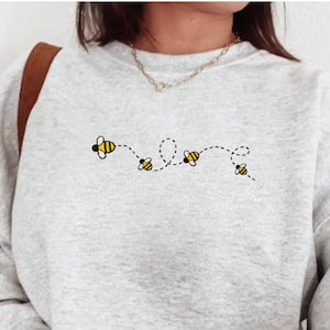 May include: A light gray sweatshirt with an embroidered design of four yellow and black bees following a dotted line. The word "EMBROIDERED" is printed in a black rectangle at the bottom.