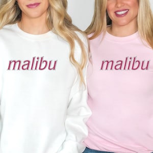 May include: Two women wearing matching sweatshirts with the word "malibu" printed in pink on the front. One sweatshirt is white and the other is pink. Both women are wearing blue jeans.