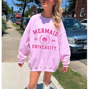 May include: Pink sweatshirt with a graphic print that says "Mermaid University Est. 1989" with a seashell and laurel wreath design. The model is wearing white cowboy boots.