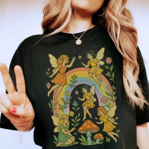 May include: Black t-shirt featuring a whimsical design of fairies, a rainbow, and mushrooms. The fairies are depicted in various poses, with colorful wings and dresses. The design is surrounded by flowers and foliage, creating a magical, nature-inspired aesthetic.