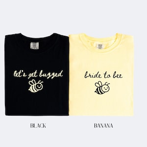 May include: Two t-shirts, one black and one banana-colored, with bee-themed graphics. The black shirt says "let's get buzzed" and the banana shirt says "bride to bee". Both shirts have a cartoon bee illustration.