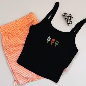 May include: A black tank top with three embroidered Halloween-themed popsicle designs, paired with orange striped shorts and a checkered hair clip. The shorts and tank top are made of a soft material.