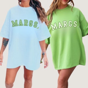 May include: Two women wearing oversized t-shirts with the word "MARGS" printed in white with a green lime slice graphic. The t-shirt on the left is light blue and the t-shirt on the right is green.