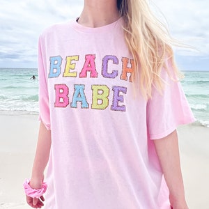 May include: A pink oversized t-shirt with the text "BEACH BABE" in rainbow colors. The shirt is worn by a person on a beach.