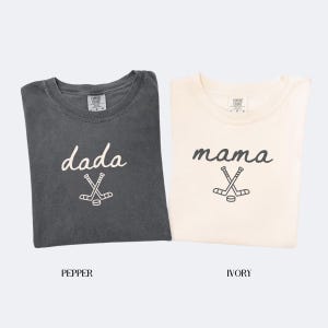 May include: Two t-shirts, one pepper-colored and one ivory-colored, are displayed. The pepper shirt has the word "dada" and a hockey stick graphic. The ivory shirt has the word "mama" and a hockey stick graphic. Both shirts are short-sleeved.