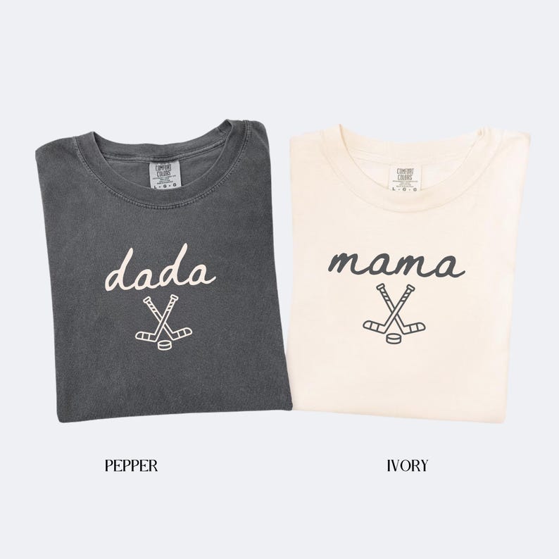 May include: Two t-shirts, one pepper-colored and one ivory-colored, are displayed. The pepper shirt has the word "dada" and a hockey stick graphic. The ivory shirt has the word "mama" and a hockey stick graphic. Both shirts are short-sleeved.