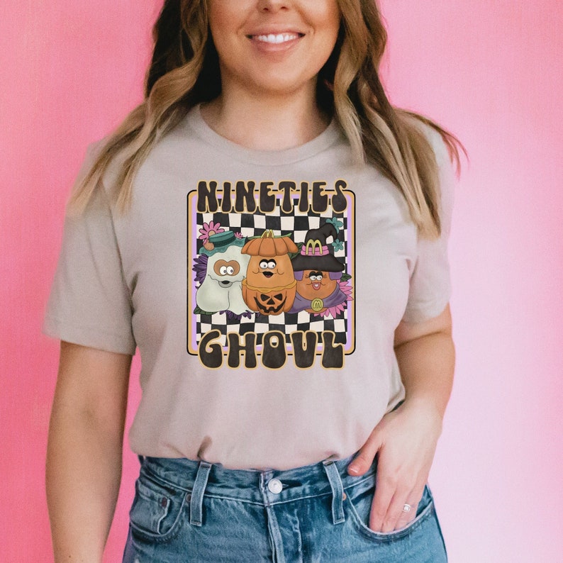 May include: A beige t-shirt featuring a graphic print of three cartoon characters dressed in Halloween costumes. The characters are a ghost, a pumpkin, and a witch. The text "NINETIES GHOUL" is printed in brown letters on the shirt.