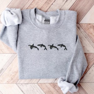 May include: A light gray sweatshirt featuring a row of four black and white orca whale illustrations. The sweatshirt has a crew neck and long sleeves. The garment is made of a soft material.