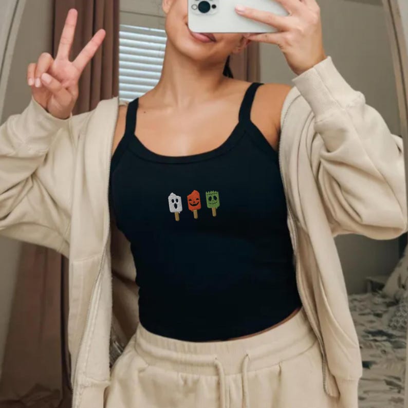May include: Black tank top with three embroidered Halloween-themed popsicle designs in white, orange, and green. The tank top has thin straps and a ribbed texture. The person is wearing a beige zip-up hoodie and beige sweatpants.