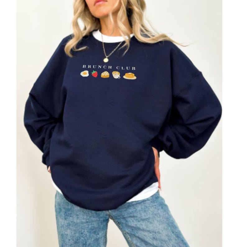 Pu&ograve; includere: Felpa blu navy con la scritta "BRUNCH CLUB" sopra icone di cibo ricamate. Le icone includono un uovo, una fragola, un pasticcino, una tazza di caff&egrave; e un pancake. La felpa ha uno scollo tondo bianco.