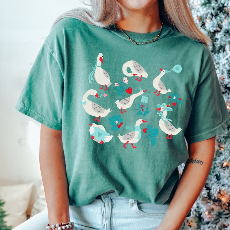 May include: A green t-shirt with a graphic print of white geese with red and blue accents. The geese are surrounded by green leaves and flowers.