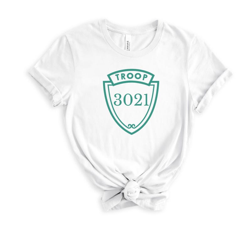 May include: White t-shirt with a teal shield design. The shield has the word "TROOP" at the top and the number "3021" in the center. The shirt is made of a soft material and has a knot at the bottom.