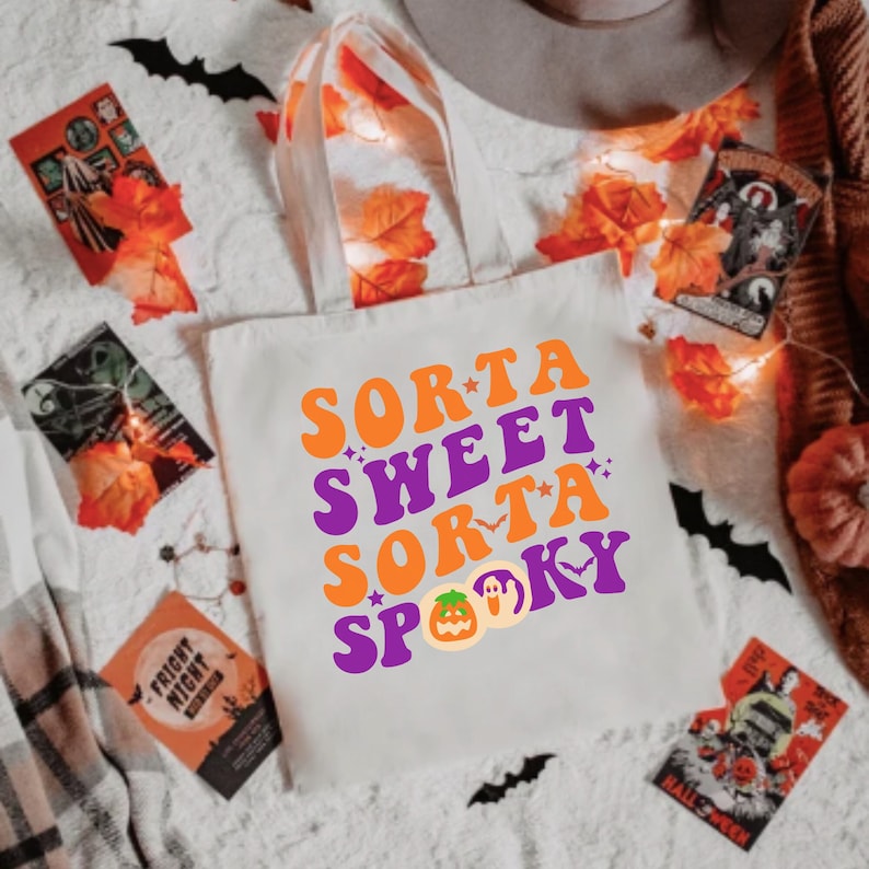 May include: A white canvas tote bag with the text "Sorta Sweet Sorta Spooky" in orange and purple letters. The text is in a retro font style. There is a pumpkin and a ghost graphic below the text.