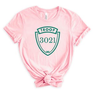 May include: A light pink t-shirt with a green shield design. The shield has the word "TROOP" at the top and the number "3021" in the center. The shirt is knotted at the bottom.