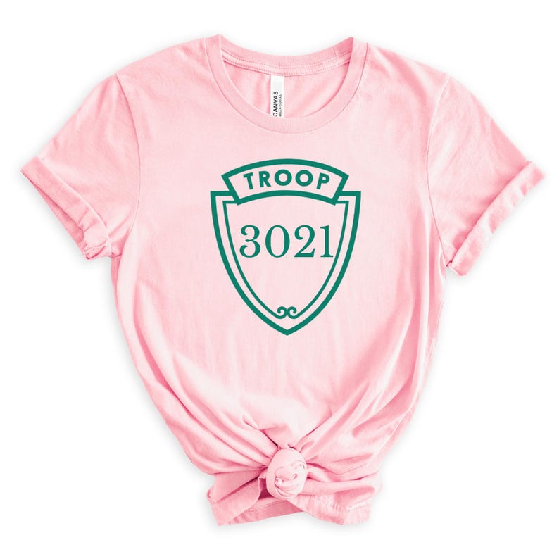 May include: A light pink t-shirt with a green shield design. The shield has the word "TROOP" at the top and the number "3021" in the center. The shirt is knotted at the bottom.