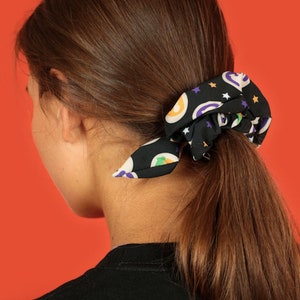 Pode incluir: Um scrunchie preto com uma estampa colorida de estrelas, ab&oacute;boras e fantasmas. O scrunchie est&aacute; amarrado em um rabo de cavalo na cabe&ccedil;a de uma pessoa.