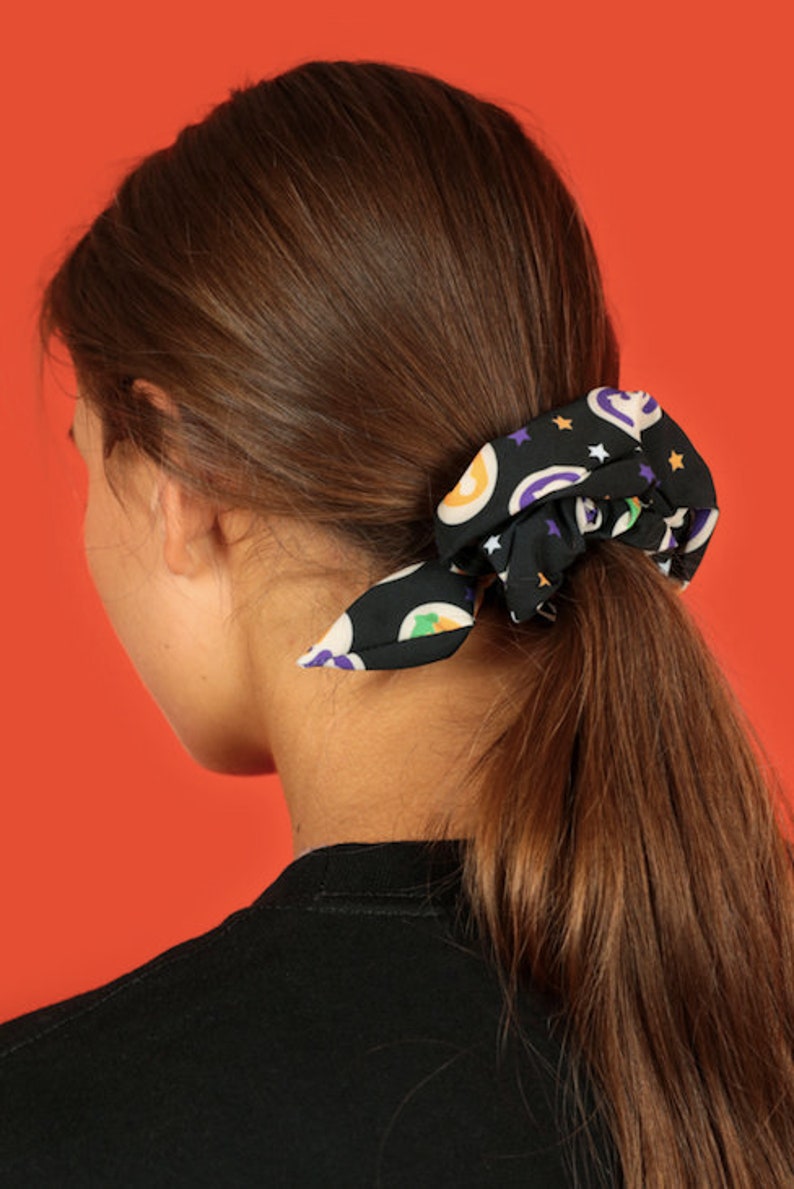 Pode incluir: Um scrunchie preto com uma estampa colorida de estrelas, ab&oacute;boras e fantasmas. O scrunchie est&aacute; amarrado em um rabo de cavalo na cabe&ccedil;a de uma pessoa.