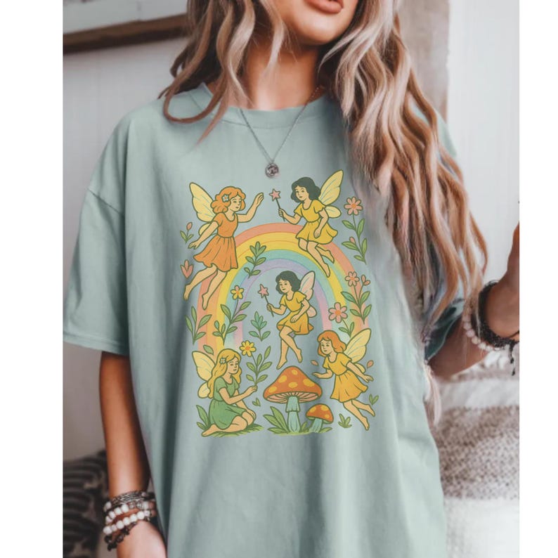 May include: A light blue t-shirt featuring a whimsical illustration of fairies, a rainbow, and mushrooms. The fairies are depicted in various poses, with colorful dresses and wings. The design has a vintage, retro aesthetic.