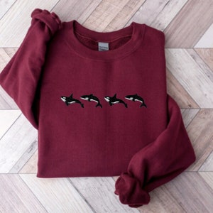 May include: A maroon sweatshirt featuring four embroidered orca whales in black and white. The orcas are horizontally aligned across the chest of the sweatshirt. The long sleeves are cuffed at the wrists.