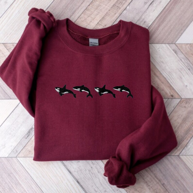May include: A maroon sweatshirt featuring four embroidered orca whales in black and white. The orcas are horizontally aligned across the chest of the sweatshirt. The long sleeves are cuffed at the wrists.