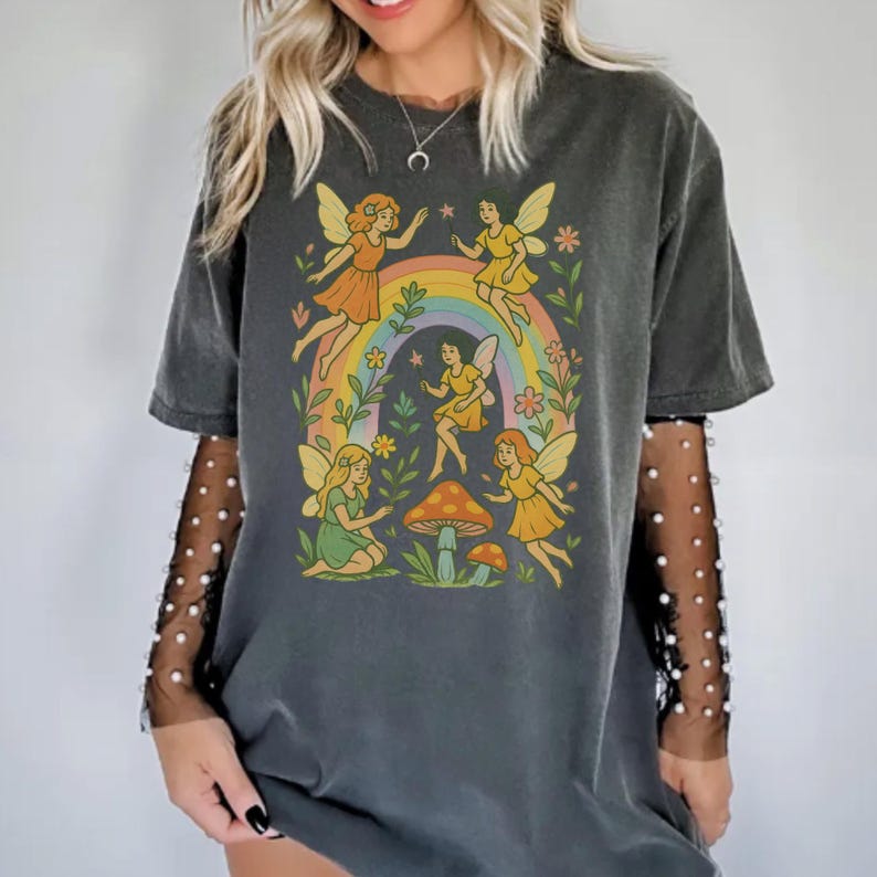 May include: Dark gray t-shirt featuring a whimsical illustration of fairies around a rainbow, mushrooms, and flowers. The fairies are depicted in yellow and orange dresses with butterfly wings. The shirt has a vintage, washed look.