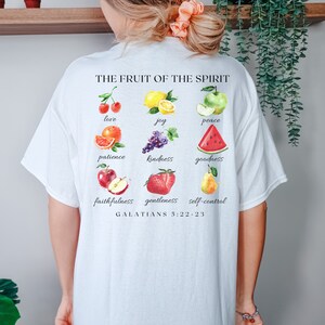 May include: White t-shirt featuring a graphic of fruit and the text "THE FRUIT OF THE SPIRIT" and "GALATIANS 5:22-23". The fruit includes cherries, a lemon, an apple, an orange, grapes, a watermelon, a strawberry, and a pear.
