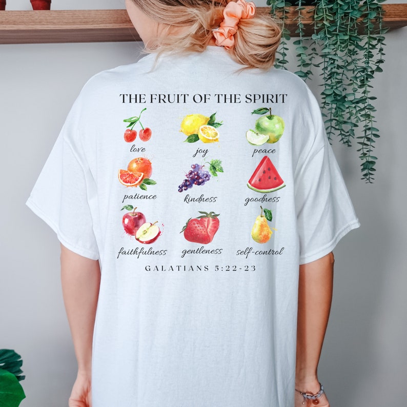 May include: White t-shirt featuring a graphic of fruit and the text "THE FRUIT OF THE SPIRIT" and "GALATIANS 5:22-23". The fruit includes cherries, a lemon, an apple, an orange, grapes, a watermelon, a strawberry, and a pear.