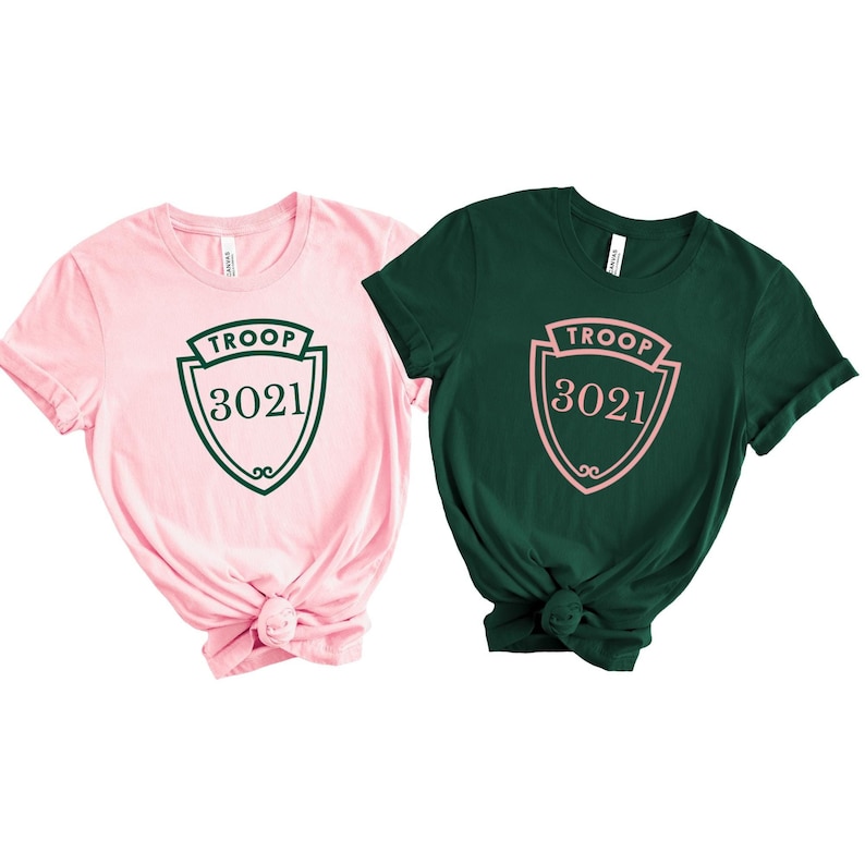 May include: Two short-sleeved t-shirts, one pink and one dark green. Each shirt features a shield design with the text "TROOP 3021" in a contrasting color. The pink shirt has a dark green design, and the green shirt has a pink design.