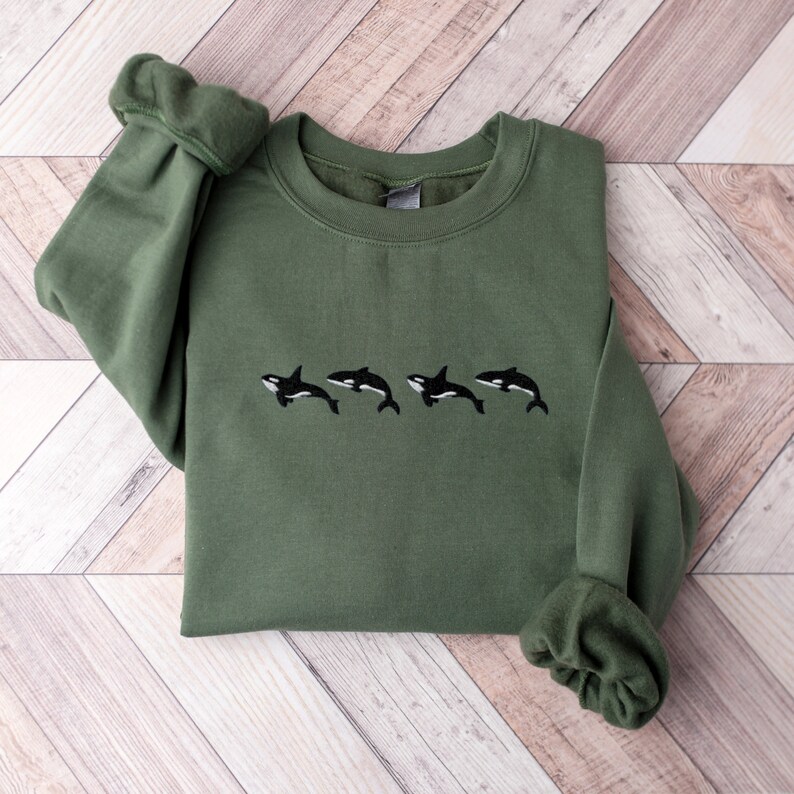 May include: An olive green sweatshirt featuring an embroidered design of four black and white orcas swimming in a row. The sweatshirt has long sleeves and a crew neck. The fabric appears soft and comfortable.