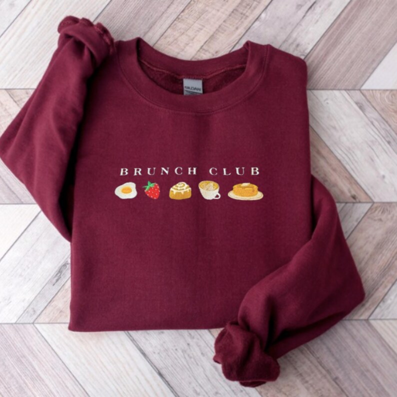 Pu&ograve; includere: Una felpa bordeaux con la scritta ricamata "BRUNCH CLUB" sopra un uovo fritto, una fragola, un rotolo alla cannella, una tazza di caff&egrave; e un pancake. Il top a maniche lunghe &egrave; realizzato in materiale morbido.