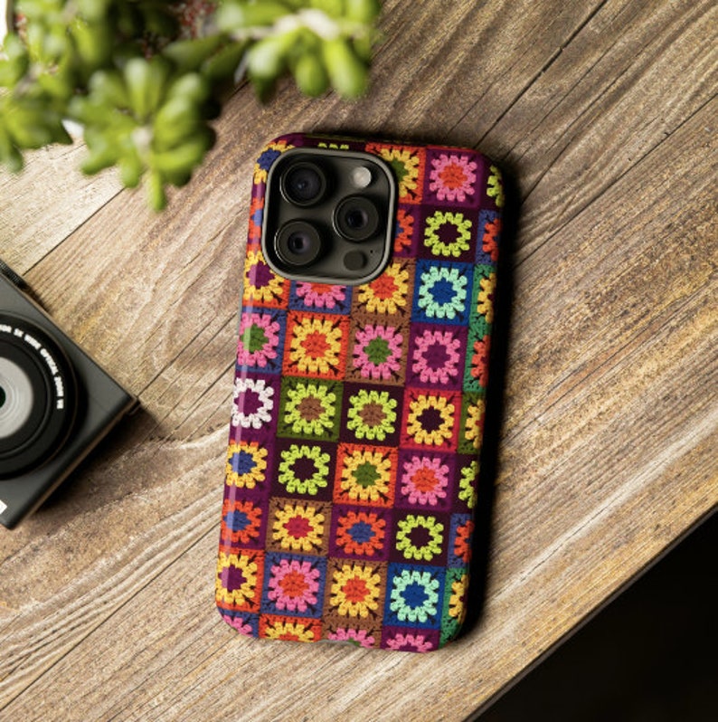 May include: A colourful phone case with a crocheted granny square pattern in shades of pink, green, yellow, blue, and brown. The case has a cutout for the phone's camera.