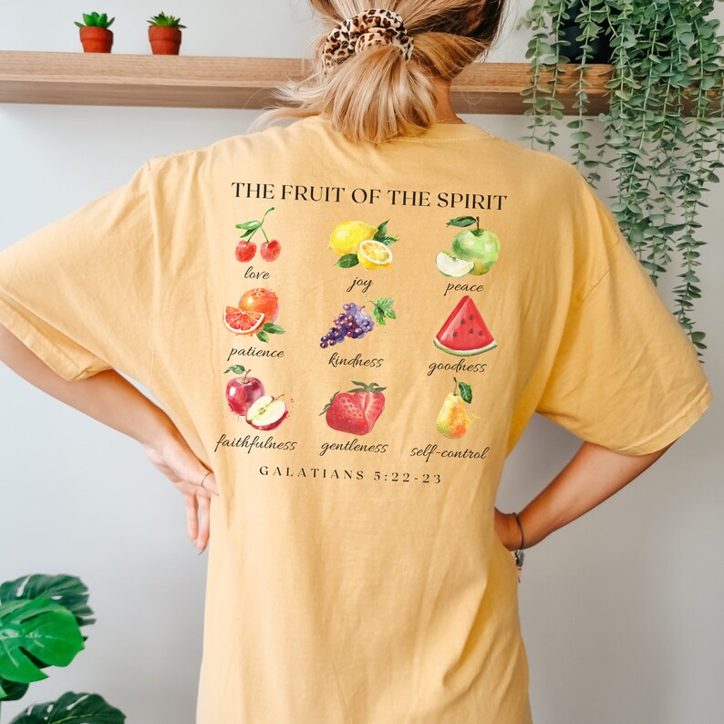 May include: A yellow t-shirt featuring a graphic of fruit and the text "THE FRUIT OF THE SPIRIT" and "GALATIANS 5:22-23". The fruit includes a cherry, lemon, apple, orange, grapes, watermelon, apple, strawberry, and pear.