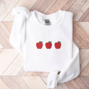May include: White sweatshirt with three red embroidered apples on the front.