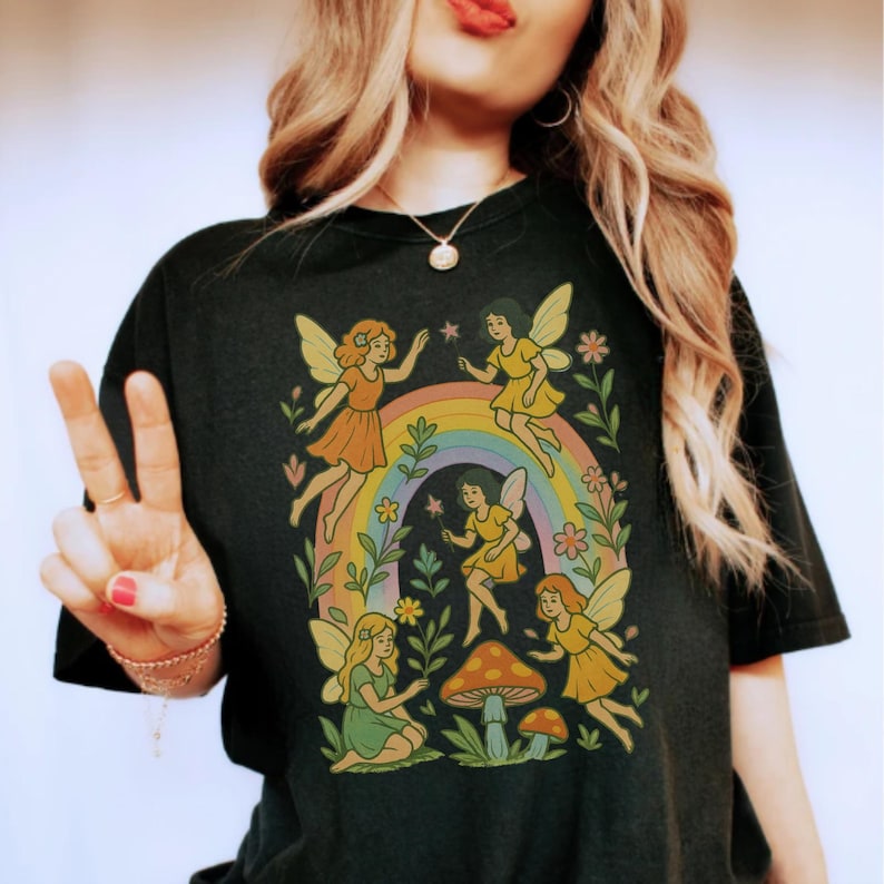 May include: Black t-shirt featuring a whimsical design of fairies, a rainbow, and mushrooms. The fairies are depicted in various poses, with colorful wings and dresses. The design is surrounded by flowers and foliage, creating a magical, nature-inspired aesthetic.