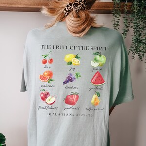 May include: A light green t-shirt featuring a graphic of fruit and the text "THE FRUIT OF THE SPIRIT" and "GALATIANS 5:22-23". The graphic includes a cherry, lemon, apple, orange, grapes, watermelon, apple, strawberry, and pear.