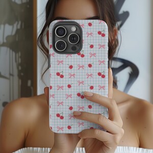 Pode incluir: Uma capa de telefone branca com um padrão xadrez azul e branco, laços rosa e cerejas vermelhas.
